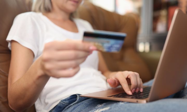 Usuaria haciendo compras online en Amazon desde su laptop. La vemos sostener su laptop en su regazo y en una mano su tarjeta de crédito.