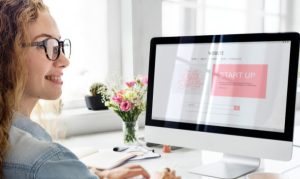 Mujer sonriente frente al monitor de una computadora, donde se aprecia que está dando apertura a su sitio web.