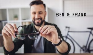 Un hombre sosteniendo unos anteojos mientras sonríe, y a un costado derecho el logo de la óptica mexicana Ben & Frank.