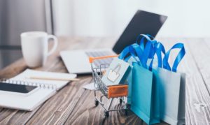 Bolsas de compras online a un costado de un carrito de compras. Al fondo encontramos una laptop, una libreta, una taza de café y un celular.