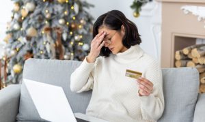 mujer tocándose la cabeza con una tarjeta de crédito en la mano, y una laptop en las piernas, sentada en una sala adornada de Navidad, representando los fraudes online durante las épocas navideñas
