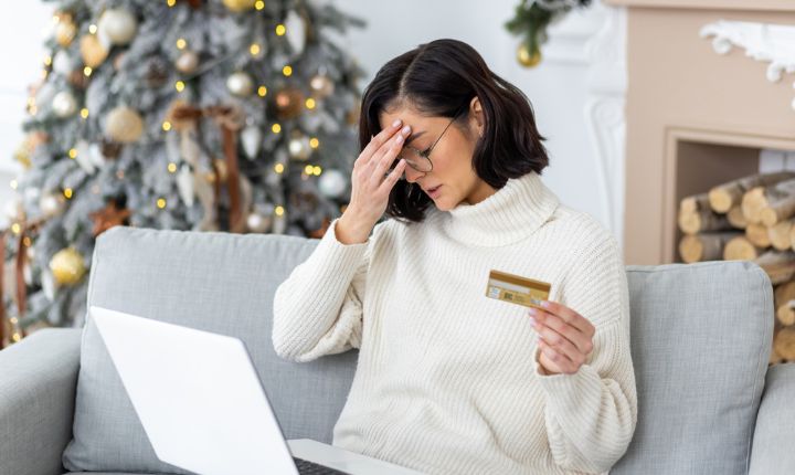 mujer tocándose la cabeza con una tarjeta de crédito en la mano, y una laptop en las piernas, sentada en una sala adornada de Navidad, representando los fraudes online durante las épocas navideñas