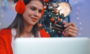 Mujer con laptop al frente, viendo los datos de su tarjeta de crédito, vestida de ropa navideña y con árbol de navidad al fondo, representando cómo compran online los mexicanos en Navidad