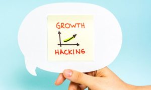 mano deteniendo un letrero blanco con un postit amarillo en el señalando como el Growth Hacking es el perfil invaluable para startups