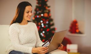 Una mujer haciendo compras online durante año nuevo a través de su laptop. La vemos sentada en su sofá, sosteniendo su laptop y su tarjeta de crédito; al fondo, un árbol de Navidad y luces decorativas.