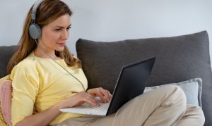 Mujer sentada en su sofá accediendo a plataformas de streaming de audio. La encontramos con su laptop en su regazo y unos audífonos.