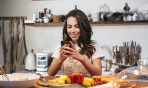 Chica en cocina con utensilios e ingredientes, mientras sostiene en una mano su celular, en el cual utiliza la app Mealstar.