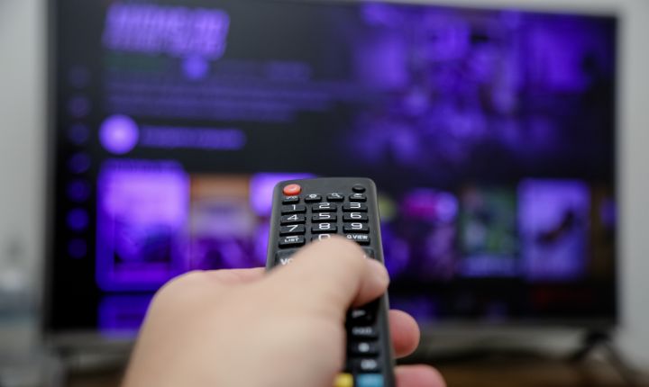 Plano subjetivo de una mano sosteniendo un control remoto, y al frente una pantalla que despliega el catálogo de una plataforma de streaming; esto en referencia al crecimiento del mercado de streaming en México.