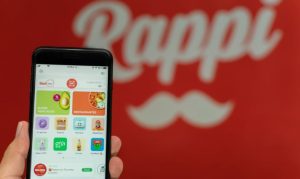 Una mano sosteniendo un celular en el que apreciamos la interfaz de Rappi, y en el fondo un back con la identidad gráfica de la app.