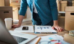 Plano medio de una empresaria recargada en un escritorio, gestionando la logística para su eCommerce y ateniendo las nuevas tendencias del mercado. En su escritorio encontramos documentos con gráficos y cifras, un vaso de café y su laptop, en tanto que en el fondo se observan paquetes de envíos.