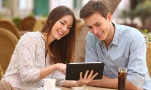 Una pareja heterosexual en México mirando videos online desde su tablet, mientras están sentados en la mesa de una cafetería y sonríen simultáneamente.